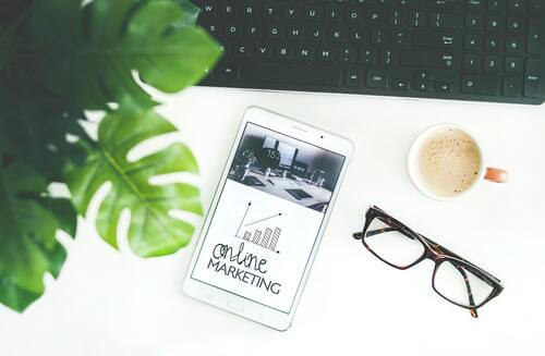 virtuelle Assistenz E-Mail-Marketing - schwarze Tastatur mit grüner Pflanze im Vordergrund und brauner Brille auf weißem Tisch und eine Kaffeetasse